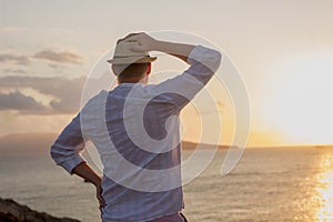 Silhouette of a man in white clothes and a hat stands with his back in the golden rays of the sun against the background of a dawn