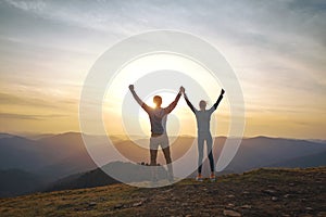 Silhouette of man and woman standing on edge of mountain and holding hands up on sunset sky and mountains background.