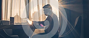 Silhouette of man at work using laptop against window background illuminated by natural sun glare