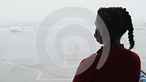 Silhouette of a Masked Young Woman at Airport Terminal Window Looking at Planes