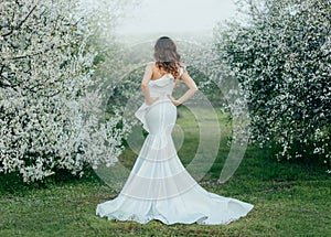 Silhouette of mysterious fantasy woman in long white elegant dress walks in green spring cherry garden. Girl runaway