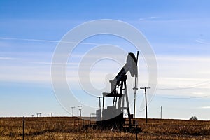 Silhouette of oil-gas pump jack out in field behind barbed wire fence silhouetted against blue sky with whispy clouds - with