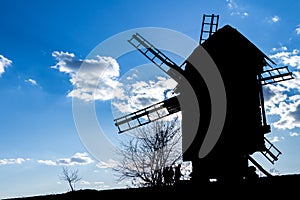 Silhouette of an old wooden mill against the background of a clear blue sky with a couple of clouds, several contours of people