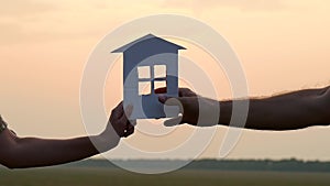 Silhouette of a paper house. The hand of a woman passes a model of a paper house in the hand of a man at sunset