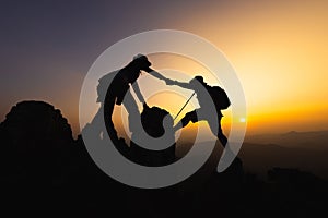 silhouette of People helping each other hike up a mountain at sunrise. Giving a helping hand, and active fit lifestyle concept.