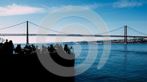 Silhouette of people relaxing in outdoor restaurant terrace overlooking the iconic 25 April bridge in Lisbon, Portugal