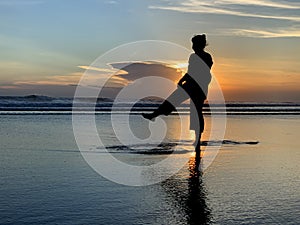 Silhouette of the person dancing alone in the beach at sunset. A woman having fun with the sea water reflection and sunlight.