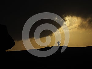 Silhouette of a person with a fishing pole standing on the hill under a yellow cloudy sky