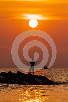 Silhouette photo of man fishing on the stone beach at sea water at the sunrise time on the morning.