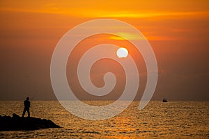 Silhouette photo of man fishing on the stone beach at sea water at the sunrise time on the morning.