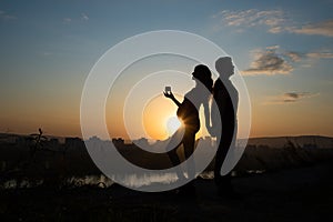 Silhouette of a pregnant girl who holds a heart in her hands and her husband on the background of a beautiful city and sunset in