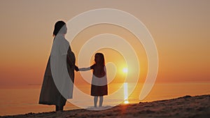 Silhouette of a pregnant woman with a baby near by. Stand near the sea at sunset. Waiting for the second change.