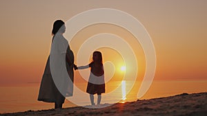 Silhouette of a pregnant woman with a baby near by. Stand near the sea at sunset. Waiting for the second change.