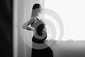 Silhouette of pregnant woman in dress at home by window touching belly. Black and white foto.