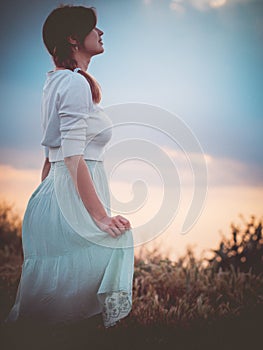 Silhouette profile of a beautiful girl in a dress in the field enjoying the sky during sunset, a young woman walking in nature,