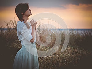 Silhouette profile of a beautiful girl in a dress praying to God in the field, a young woman walking on nature during sunset