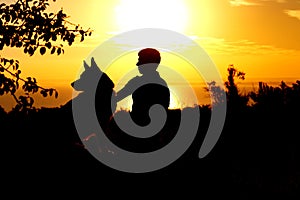 Silhouette profile of German Shepherd dog obediently sitting nearby his owner man, boy walking on nature with pet at sunset in a