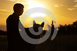Silhouette profile of a young man and a dog watching the sun set on the horizon in a field, boy fondle his pet on nature, concept photo