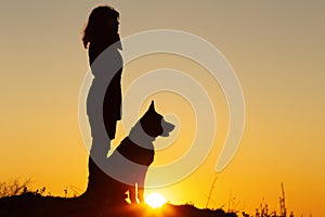 Silhouette profile of young woman and German Shepherd dog looking in the distance at sunset, pet sitting near girl`s leg on the