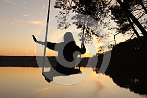 silhouette of a romantic young woman on a swing over lake at sunset. Young girl traveler sitting on the swing in