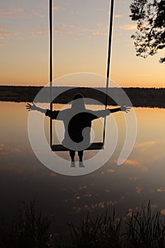 silhouette of a romantic young woman on a swing over lake at sunset. Young girl traveler sitting on the swing in