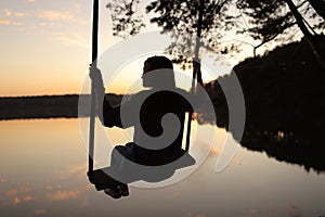 silhouette of a romantic young woman on a swing over lake at sunset. Young girl traveler sitting on the swing in