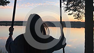 silhouette of a romantic young woman on a swing over lake at sunset. Young girl traveler sitting on the swing in