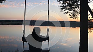 silhouette of a romantic young woman on a swing over lake at sunset. Young girl traveler sitting on the swing in