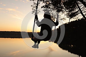 silhouette of a romantic young woman on a swing over lake at sunset. Young girl traveler sitting on the swing in