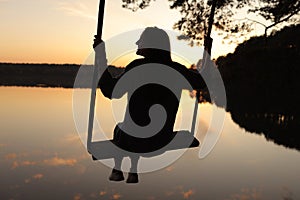 silhouette of a romantic young woman on a swing over lake at sunset. Young girl traveler sitting on the swing in