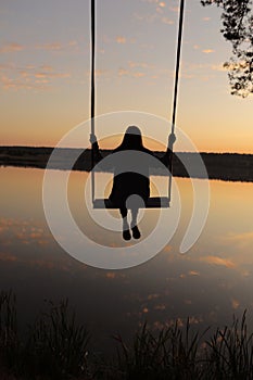 silhouette of a romantic young woman on a swing over lake at sunset. Young girl traveler sitting on the swing in