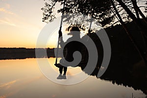 silhouette of a romantic young woman on a swing over lake at sunset. Young girl traveler sitting on the swing in