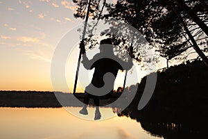 Silhouette of a romantic young woman on a swing over lake at sunset. Young girl traveler sitting on the swing in beautiful nature