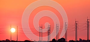 Silhouette row of electric poles with cable lines against orange sunset sky background at countryside, panoramic view