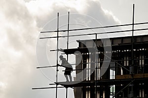 Silhouette scaffolder in PPE erecting framework of wooden planks and tall scaffolding poles high up on modern new building