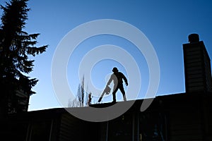 Silhouette of senior man with gas powered leaf blower cleaning roof gutters on an apartment building, fall maintenance