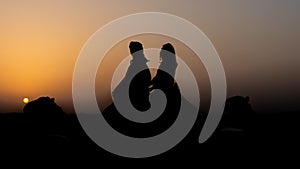Silhouette shot of two people riding a camel in Dubai desert with sunset sky on the horizon
