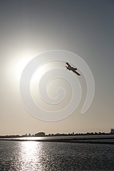 Silhouette of a single-engine aircraft with a propeller flying over  a lake with a shiny surface