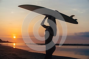 Silhouette of slim girl with surfboard in hands at beach on background of beautiful sunset