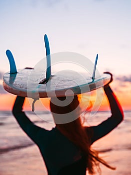 Silhouette of slim girl in wetsuit with long hair and surfboard on beach at warm sunset or sunrise. Surfer and ocean