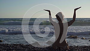 Silhouette of slim happy woman on the right raising hands sitting on pebble beach in twilight dusk. Back view confident