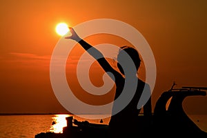 Silhouette of small girl holding sun during sunset above marina, part of motor boat visible on right side