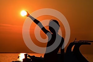 Silhouette of small girl holding sun during sunset above marina, part of motor boat visible on right side