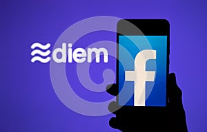 Silhouette of smartphone with Facebook on the screen and Facebook Diem crypto coin logo on the large blurred screen.