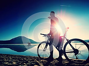 Silhouette of sportsman holding bicycle on lake beach, colorful sunset cloudy sky in background