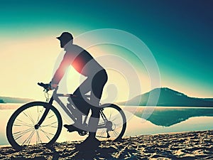 Silhouette of sportsman holding bicycle on lake beach, colorful sunset cloudy sky in background