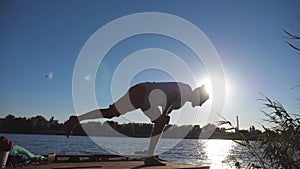 Silhouette of sporty man standing at yoga pose on a wooden jetty at lake. Young guy training at nature. Athlete