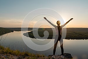 Silhouette of a sporty young woman standing on a ridge, enjoying the sunset over a river valley