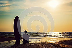 Silhouette of surf man sitting with a surfboard on the seashore