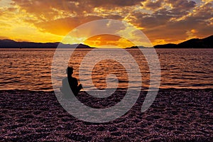 Silhouette of a teenager who sits on a pebble sea shore and watches a beautiful, vibrant sunset on the Gulf of Corinth in Greece